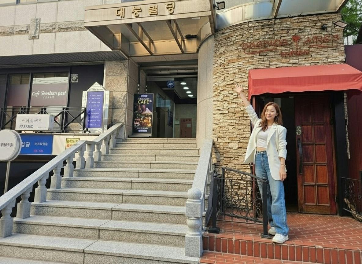 A woman stands outside a building, smiling and pointing upward. The building has a staircase and a cafe entrance.