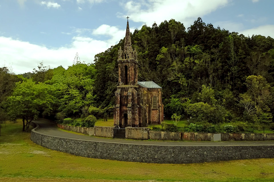 A historic chapel with a tall steeple stands in front of a dense forest, surrounded by a stone wall and greenery.