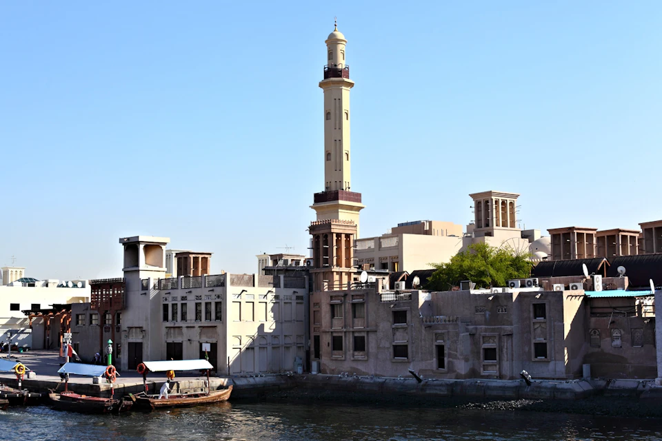 Traditional buildings and a tall minaret line a waterfront under a clear blue sky; a boat floats on the water in the foreground.