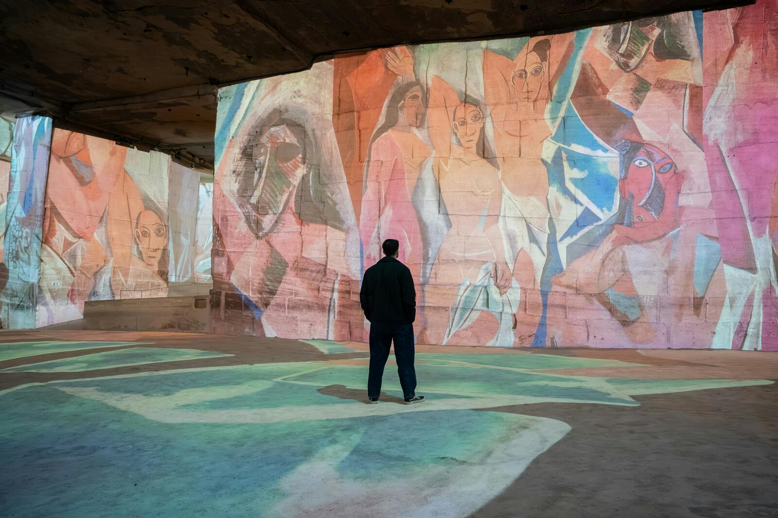 A person stands in a large room with colorful abstract art projected onto its walls and floor.
