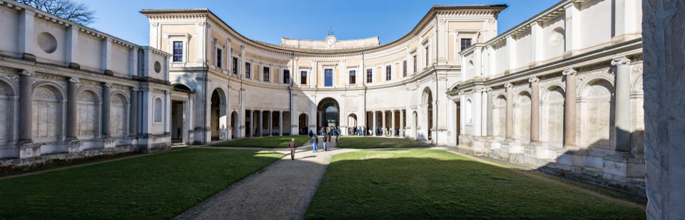People walking in a courtyard surrounded by a curved, three-story building with arched doorways and a clock above the central arch.