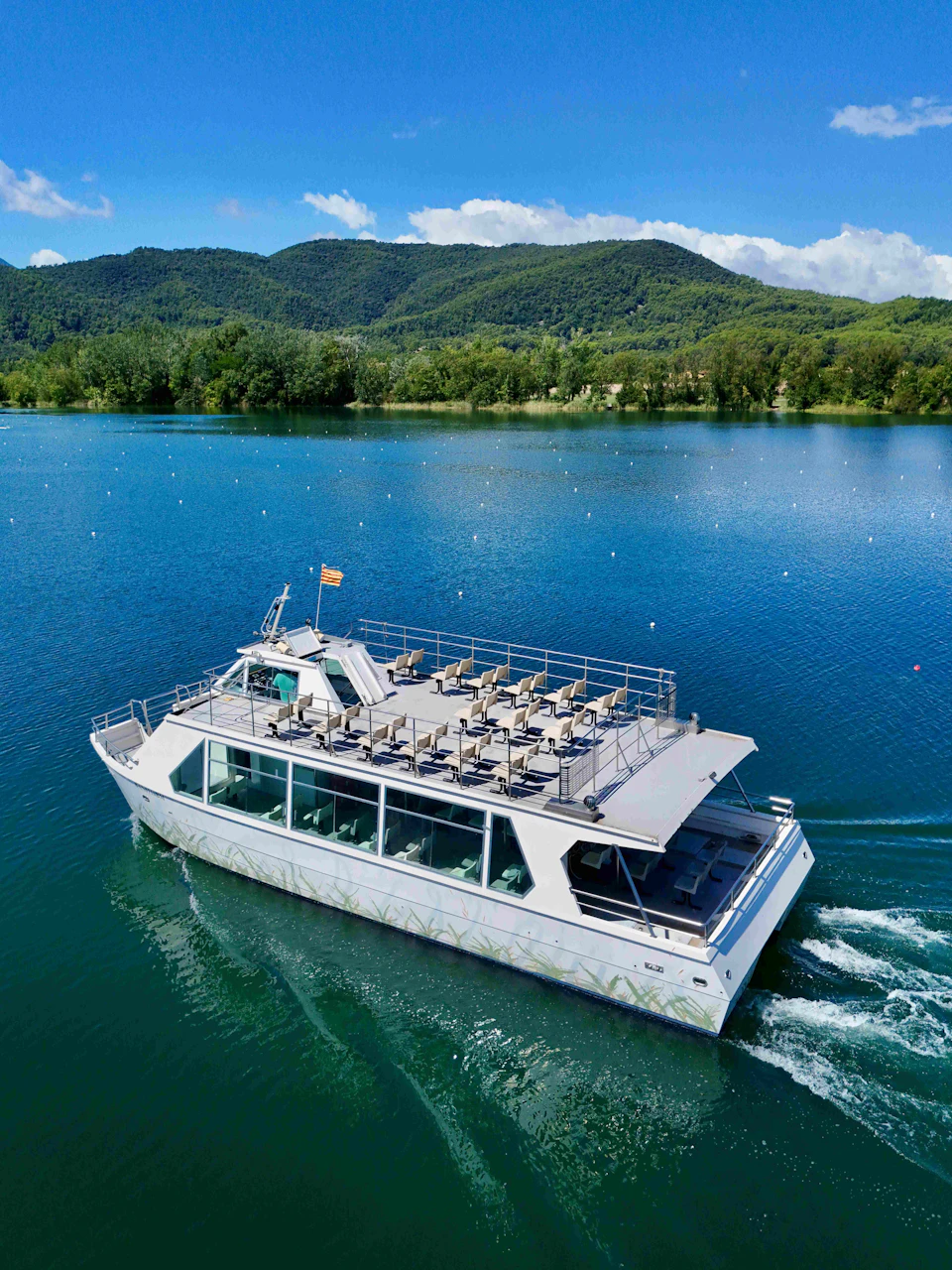 A modern boat with an upper deck of arranged chairs cruises on a clear lake, with forested hills and a partly cloudy sky in the background.