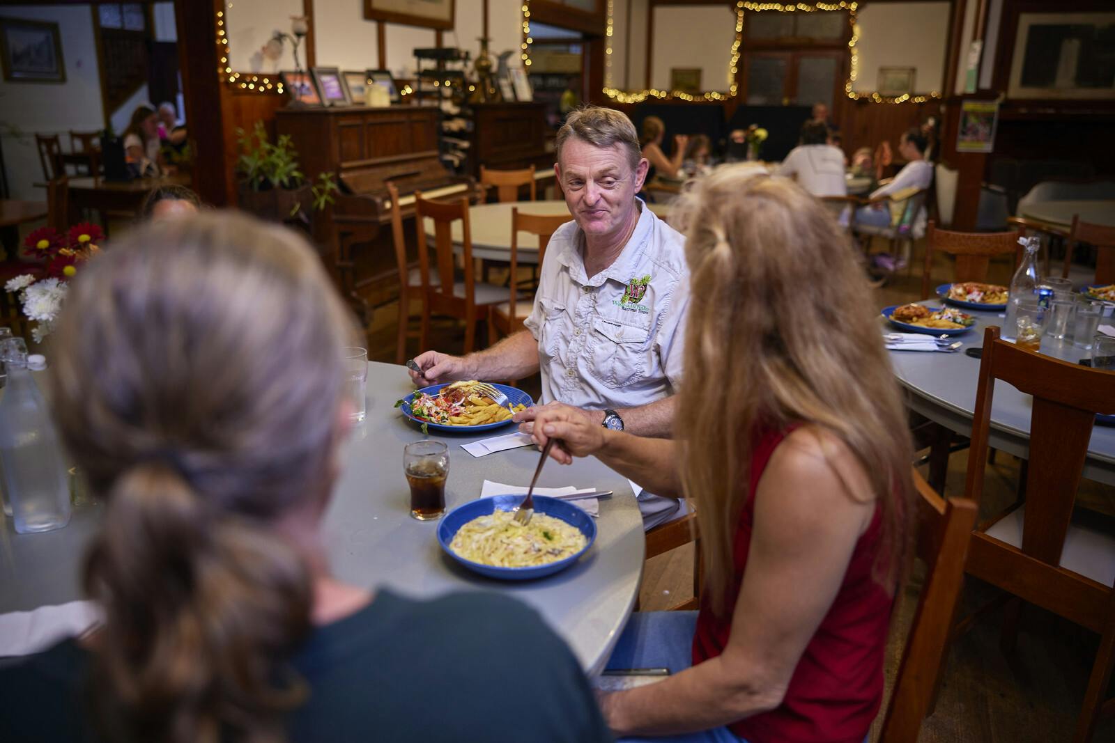 居心地の良いレストランの円卓に3人が座り、パスタを食べています。 他のダイナーはバックグラウンドで表示されます。