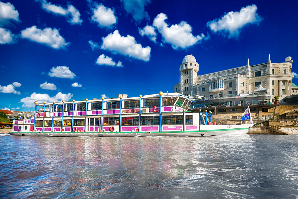 A colorful riverboat with multiple windows on a sunny day, with a historic building and a bright blue sky full of clouds in the background.