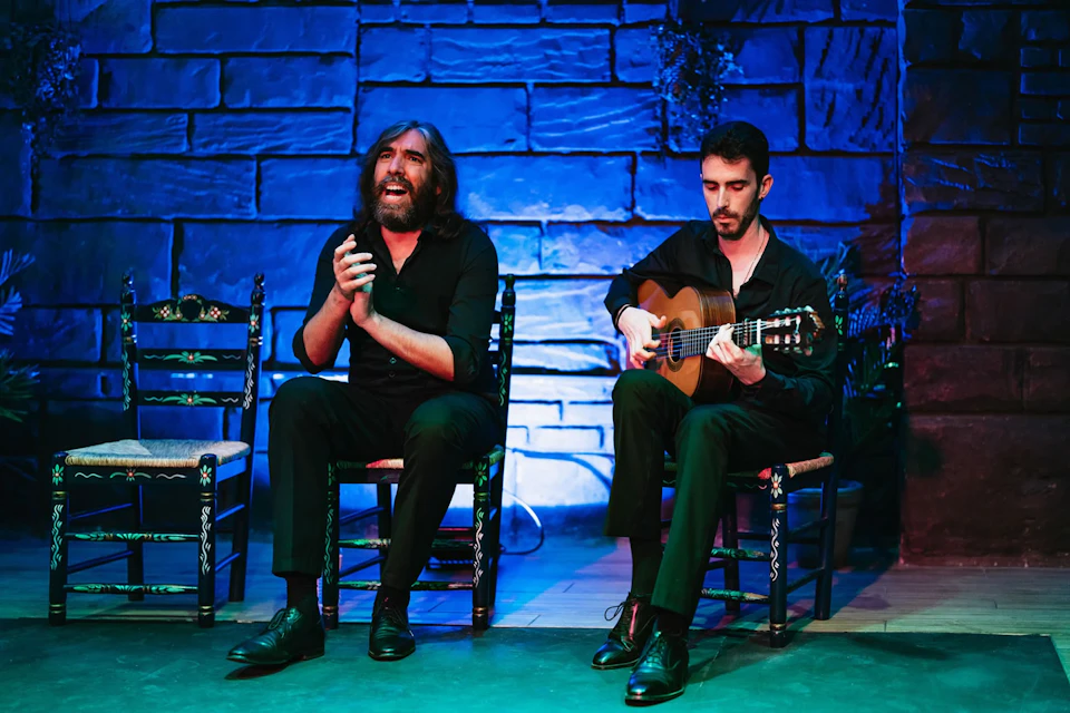 A man claps and sings while seated next to another man playing a guitar in front of a blue-lit brick wall.