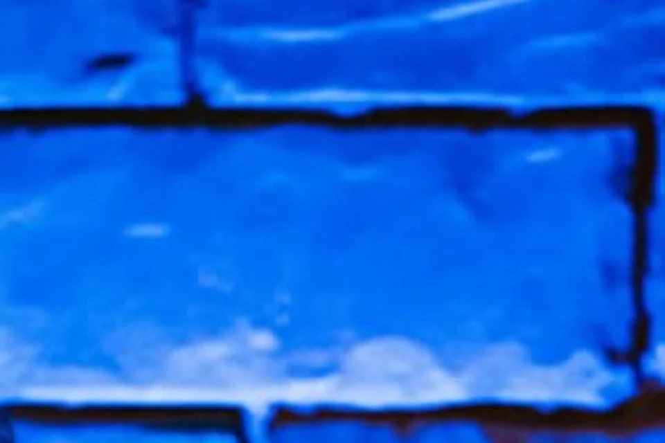 A man claps and sings while seated next to another man playing a guitar in front of a blue-lit brick wall.