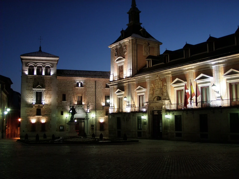 Illuminated historic building with arched windows, clock tower, and flags at dusk in a cobblestone square.