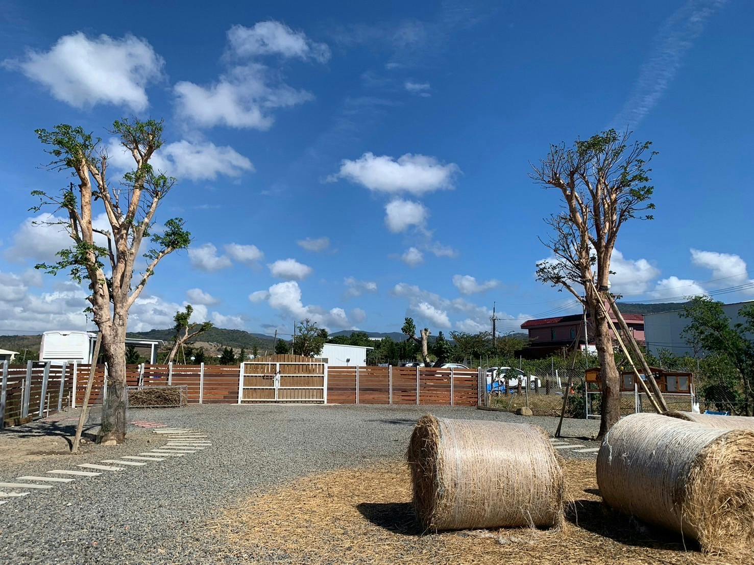 A rural scene with hay bales, trees, a fence, and buildings under a clear blue sky with scattered clouds.