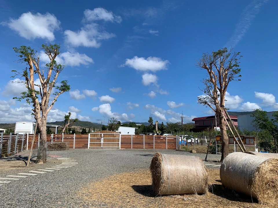 A rural scene with hay bales, trees, a fence, and buildings under a clear blue sky with scattered clouds.