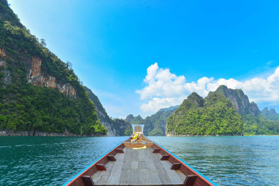 A long-tail boat travels between green, rocky hills on a blue lake under a clear sky with scattered clouds.