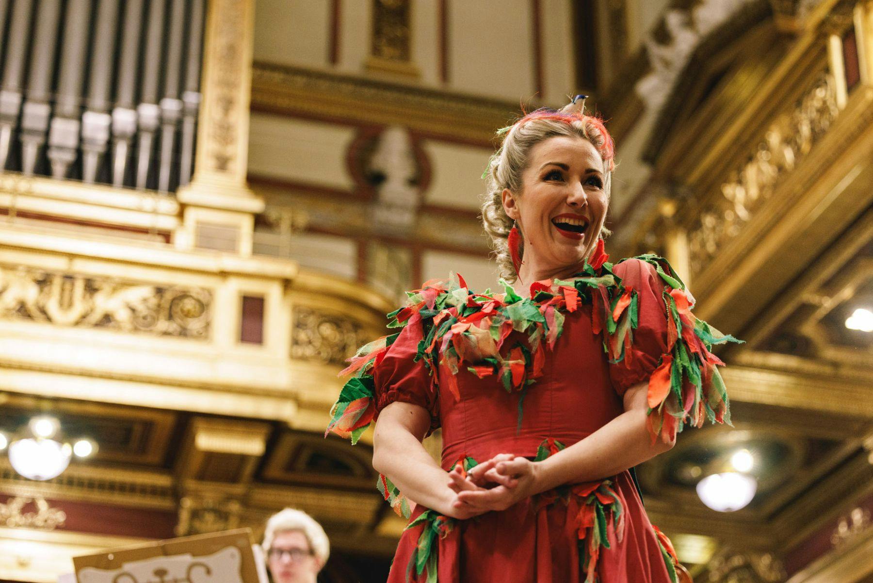 A woman in a red dress adorned with green and red leaves smiling in an ornate, gilded building interior.