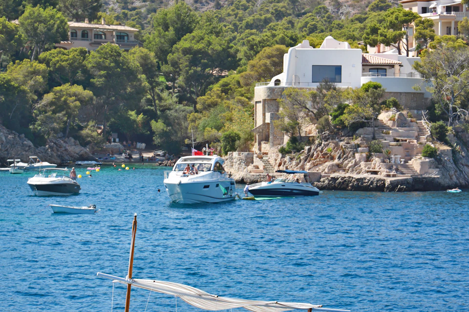Boats anchored near a rocky coastline with white buildings and lush greenery in the background.