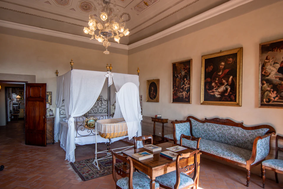 A vintage room with a canopy bed, baby cradle, wooden furniture, and classical paintings on the walls, lit by an ornate chandelier.