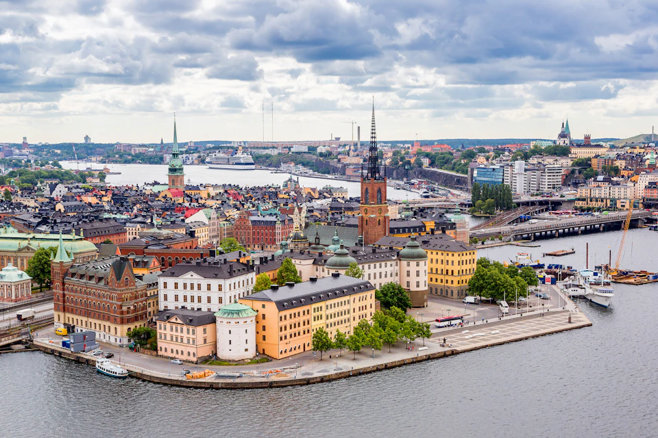 A panoramic view of a European city with colorful buildings, a prominent church tower, waterfront, and cloudy sky.