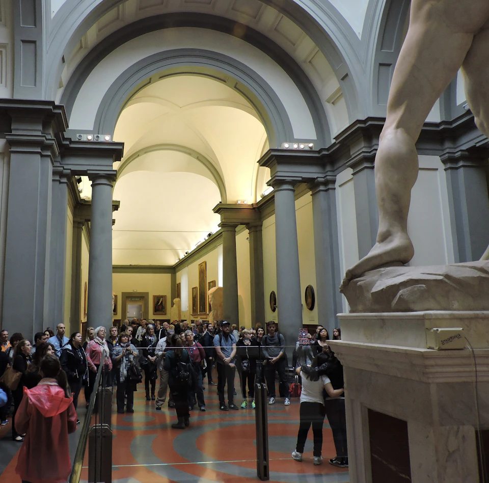A crowd of people in a museum standing around a sculpture, viewed from behind the leg of a large marble statue.