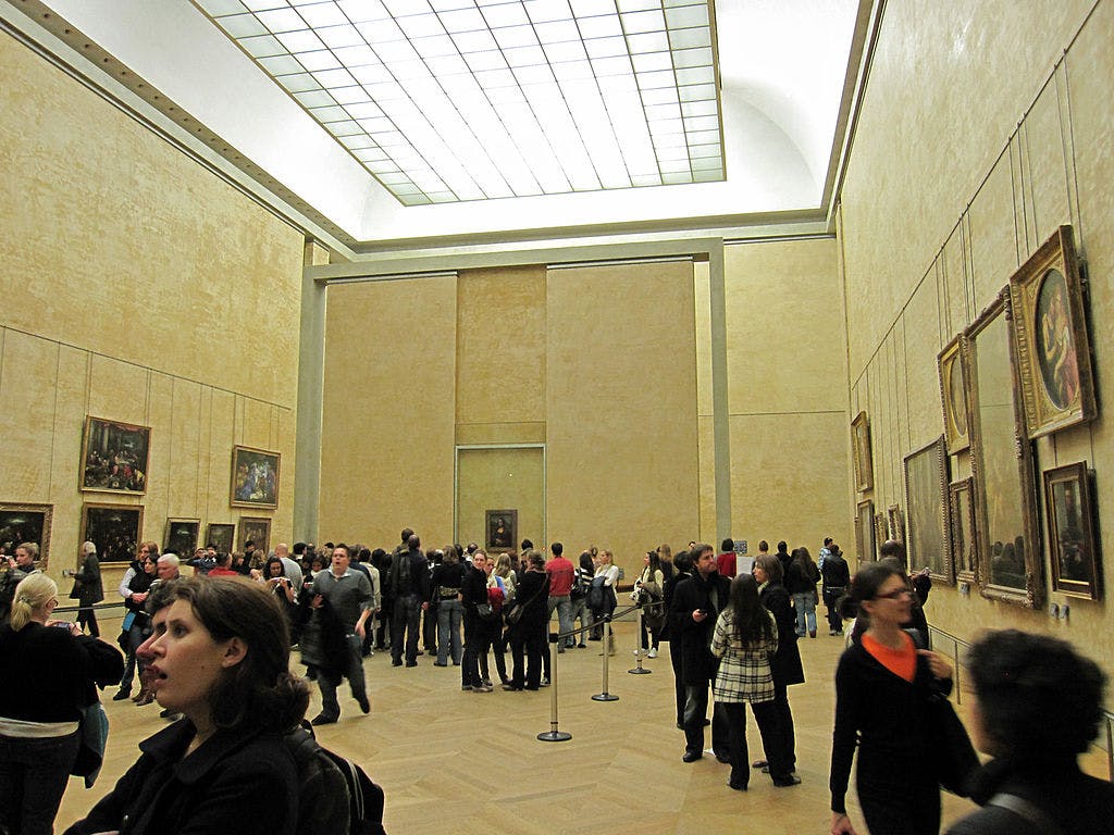 Crowded art gallery with people viewing paintings on both walls under a large, bright ceiling.
