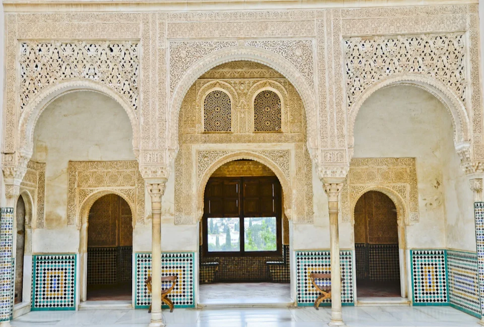 A richly decorated interior with arched doorways, intricate patterns, tiled walls, and a central window overlooking a garden.