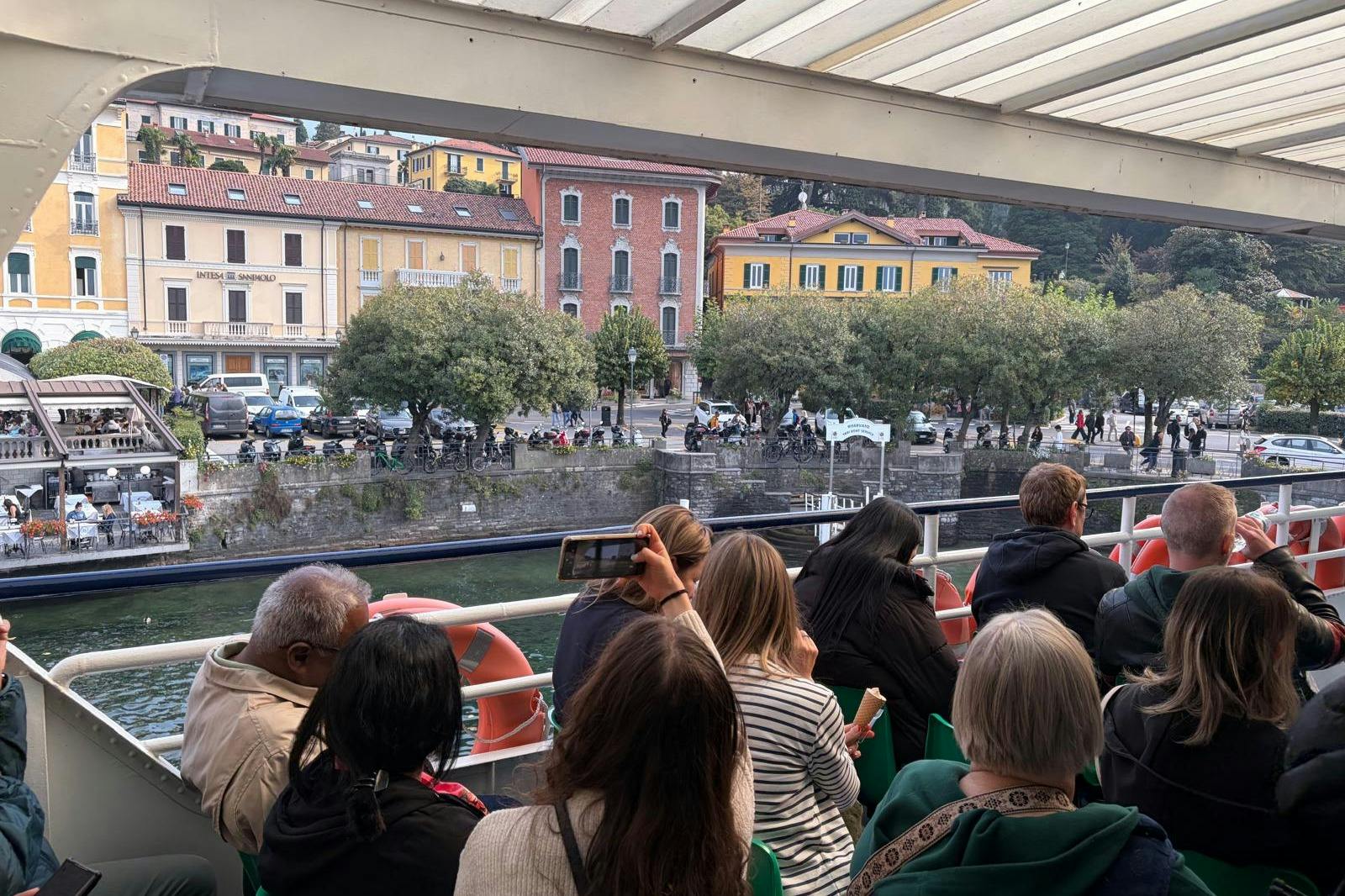 People on a boat look towards a waterfront with colorful buildings and trees. One person is taking a photo with a phone.