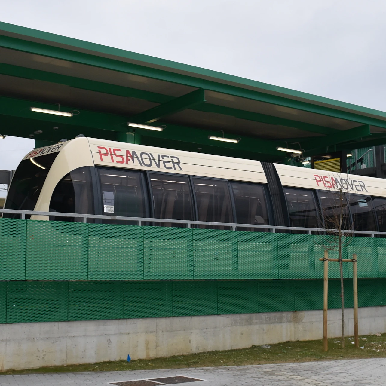 A PisaMover tram stopped at a modern green station platform on a cloudy day.