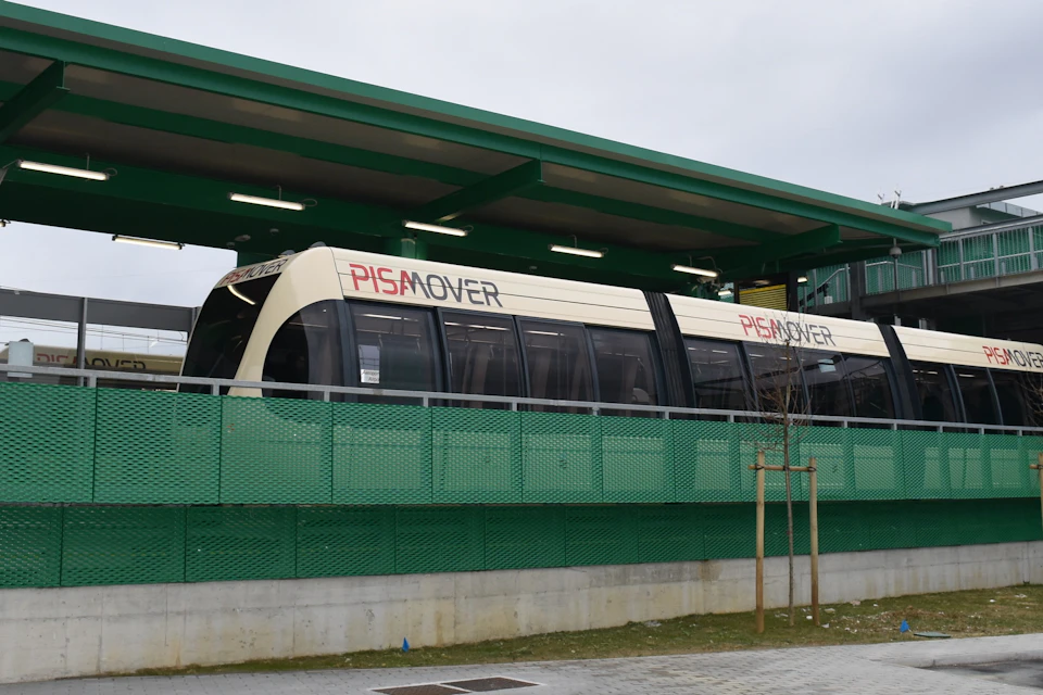 A PisaMover tram stopped at a modern green station platform on a cloudy day.