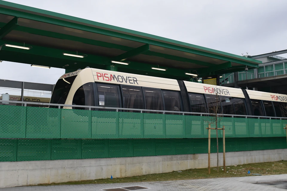 A PisaMover tram stopped at a modern green station platform on a cloudy day.