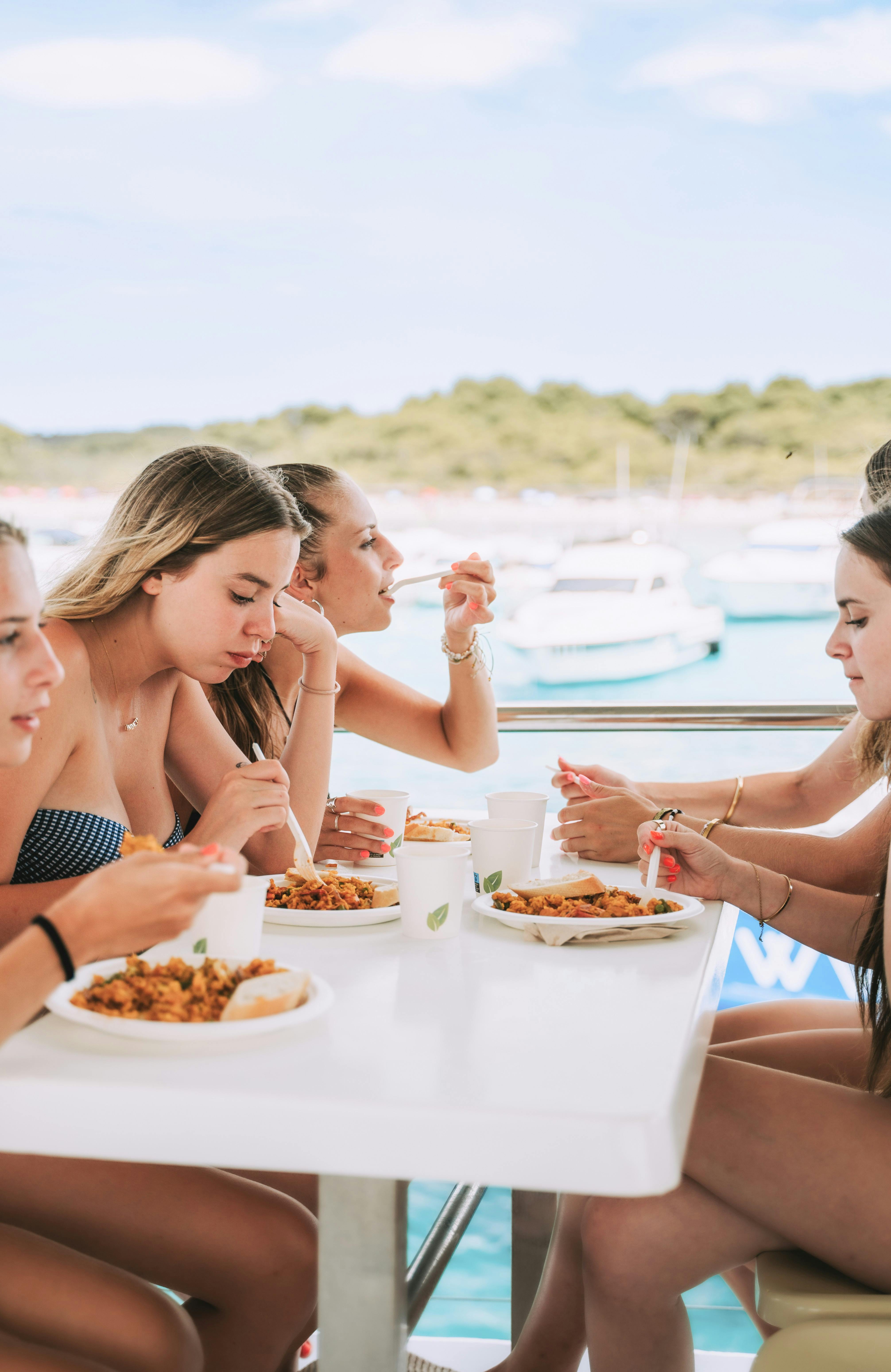 A group of people seated outdoors, eating food from plates at a table, with boats and trees visible in the background.