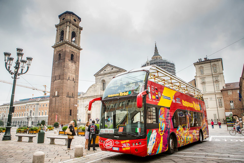 Tourists near a colorful double-decker sightseeing bus in a historic square with a tall bell tower and several old buildings.