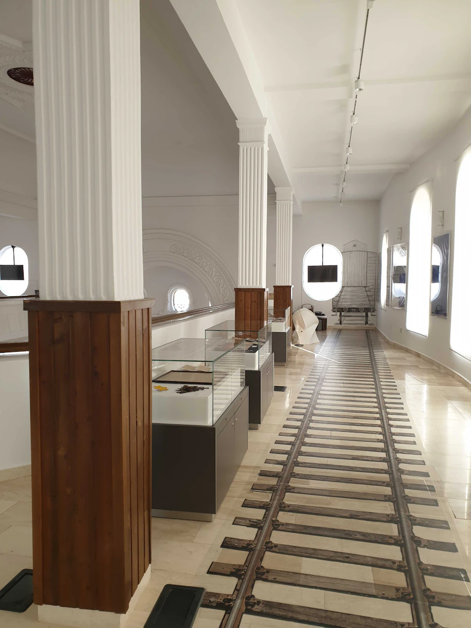 A white hall with glass display cases, wooden accents, tiled floor, and round windows allowing natural light.