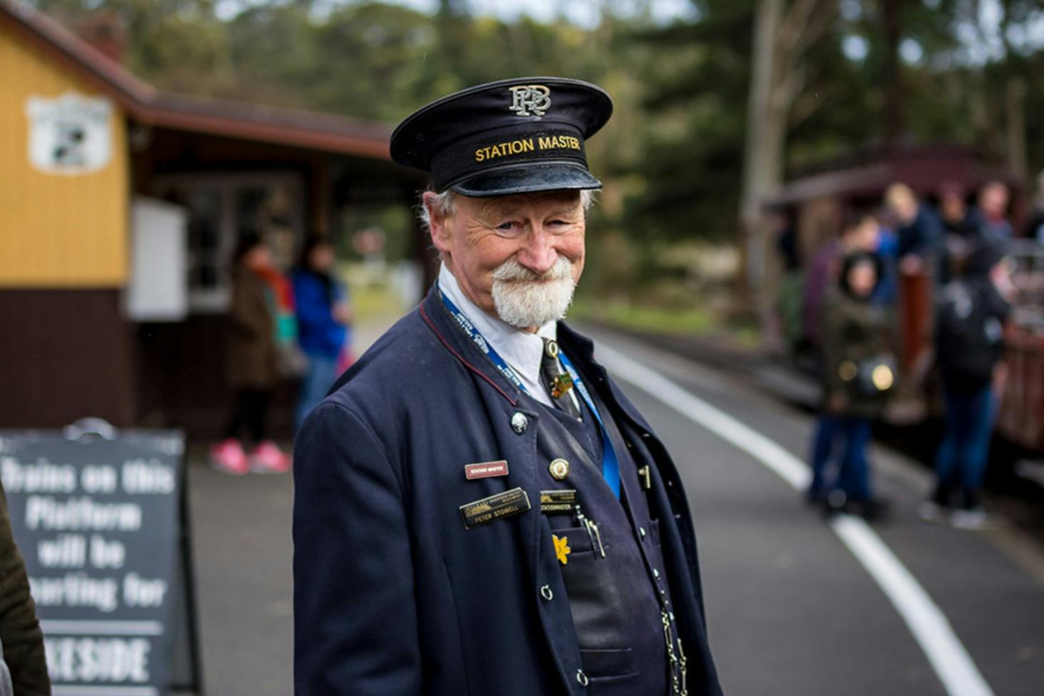 A station master in uniform smiling, standing at a railway platform with blurred people and greenery in the background.