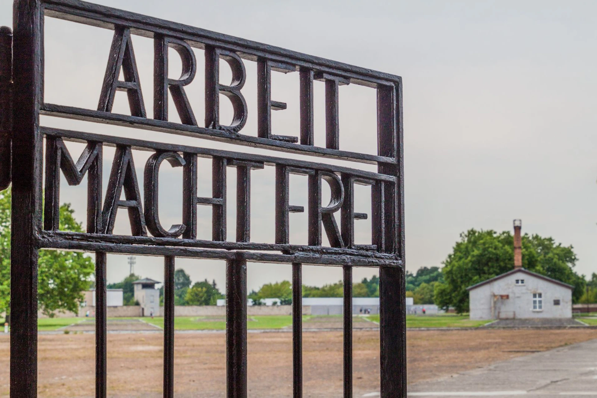 Wrought iron gate with "Arbeit Macht Frei" inscription, overlooking a barren, open area with buildings and trees in the distance.