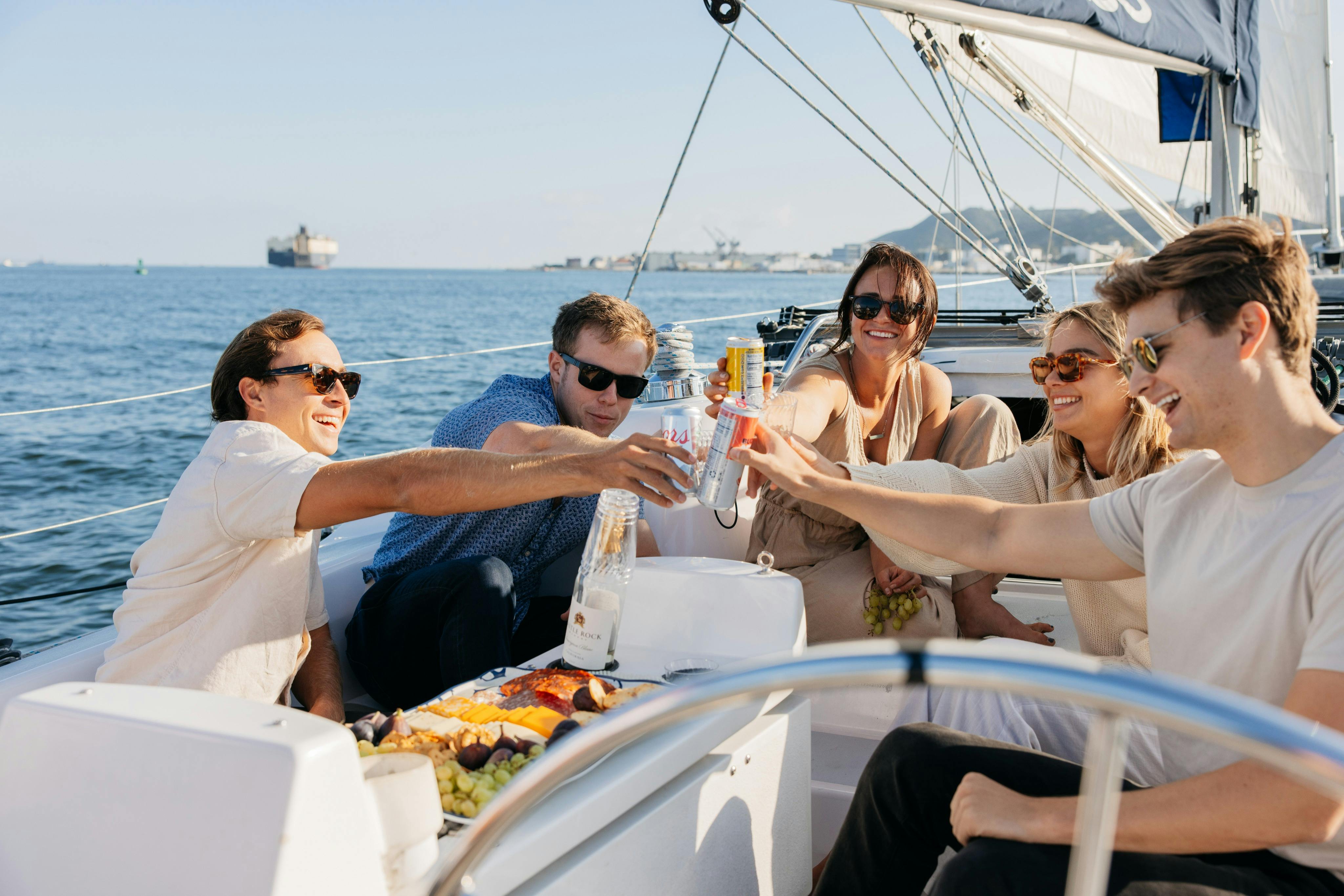 Group of young adults sitting in the cockpit of a sailboat toasting drinks with Point Loma in the background