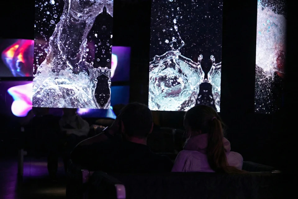 Two people sitting in a dark room with large screens displaying water splash imagery in the background.