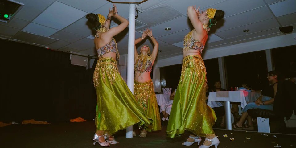 Three dancers in green skirts, adorned with gold accessories, perform in a room with tables and white tablecloths.