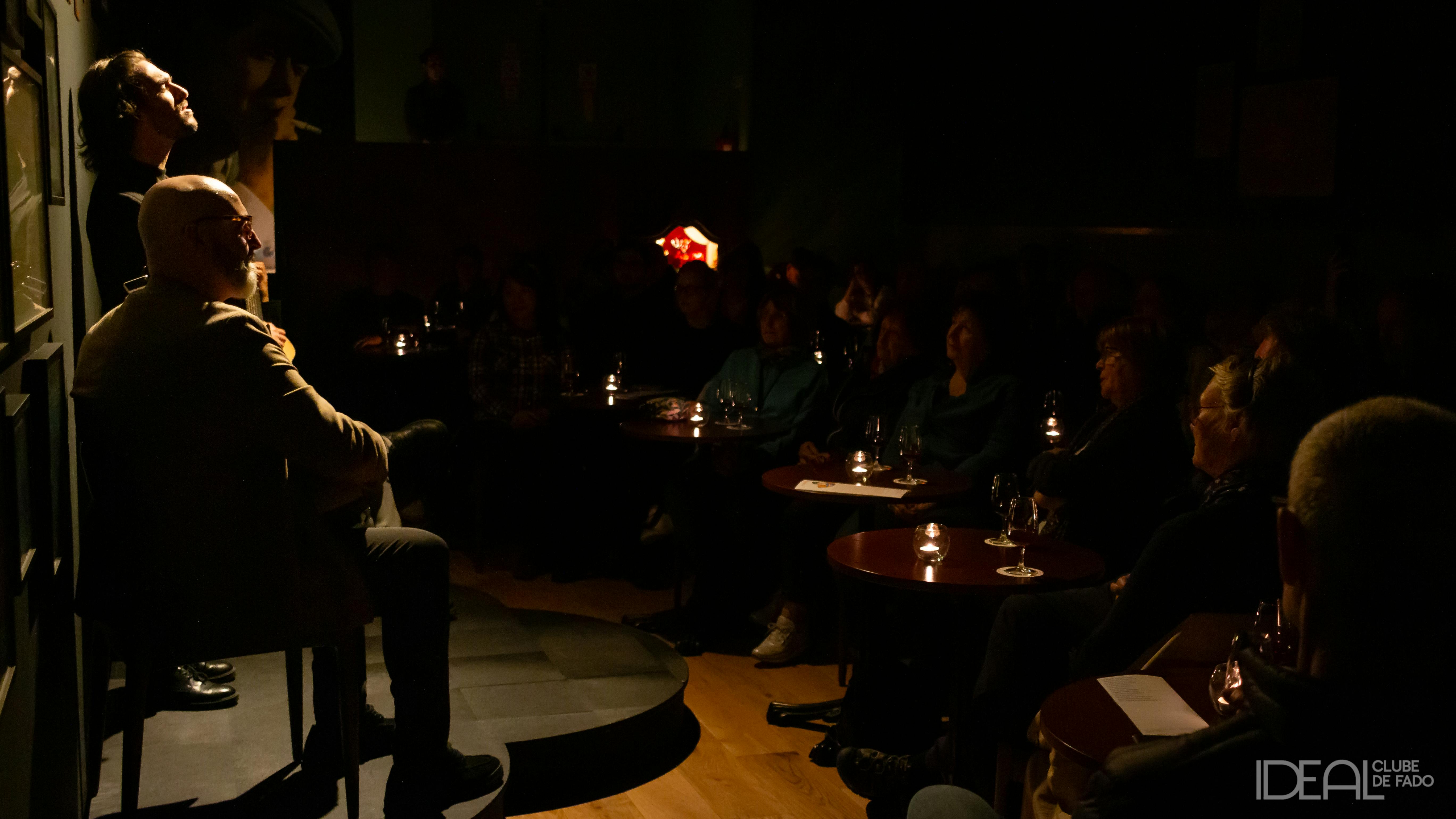 Dva hudebníci vystupují na slabě osvětleném pódiu před pozorným publikem v útulném, intimním prostředí s malými stoly a světlem svíček.