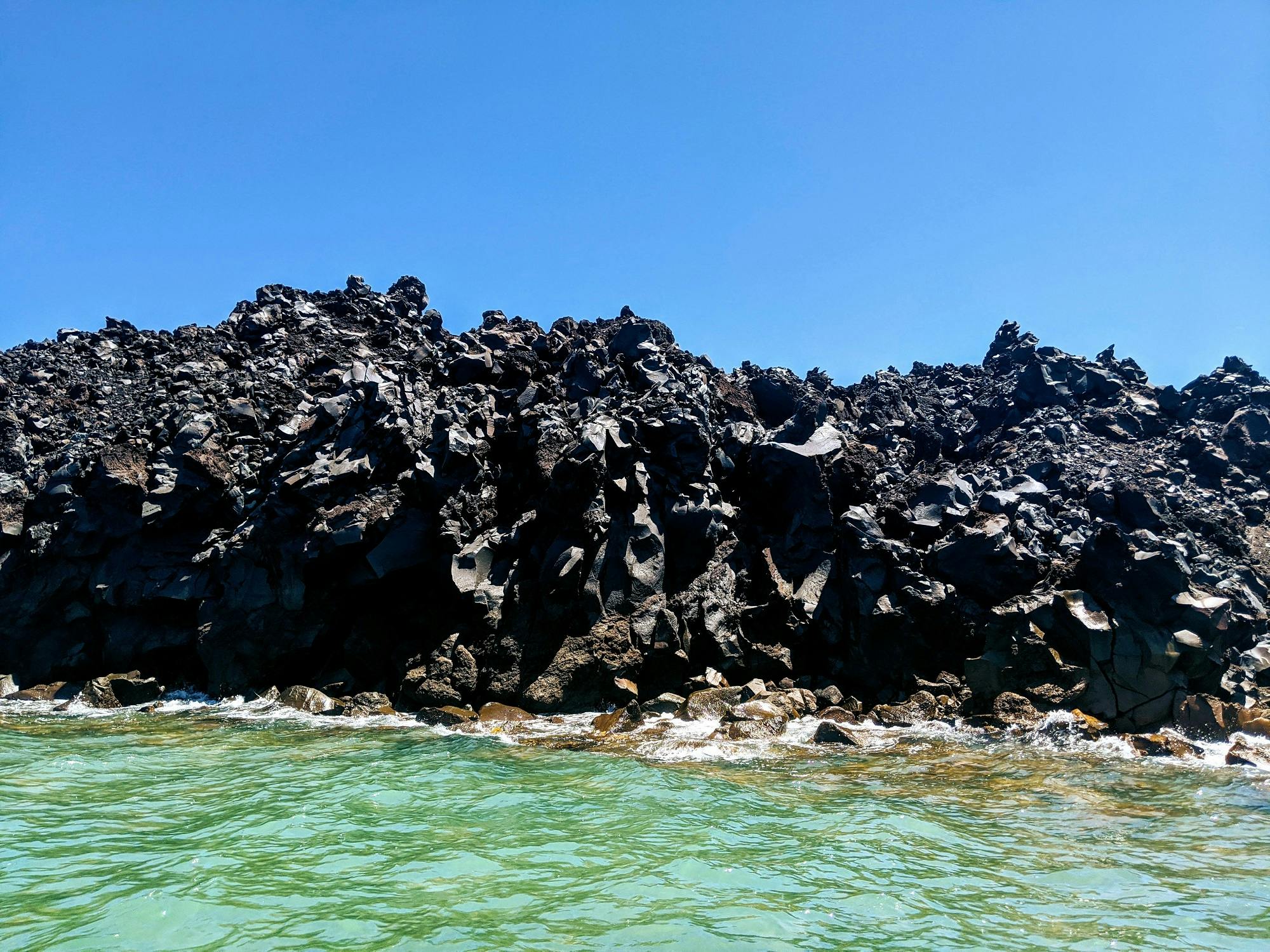 岩石黑色熔岩在晴朗的蓝天下形成，底部轻轻拍打着海浪。