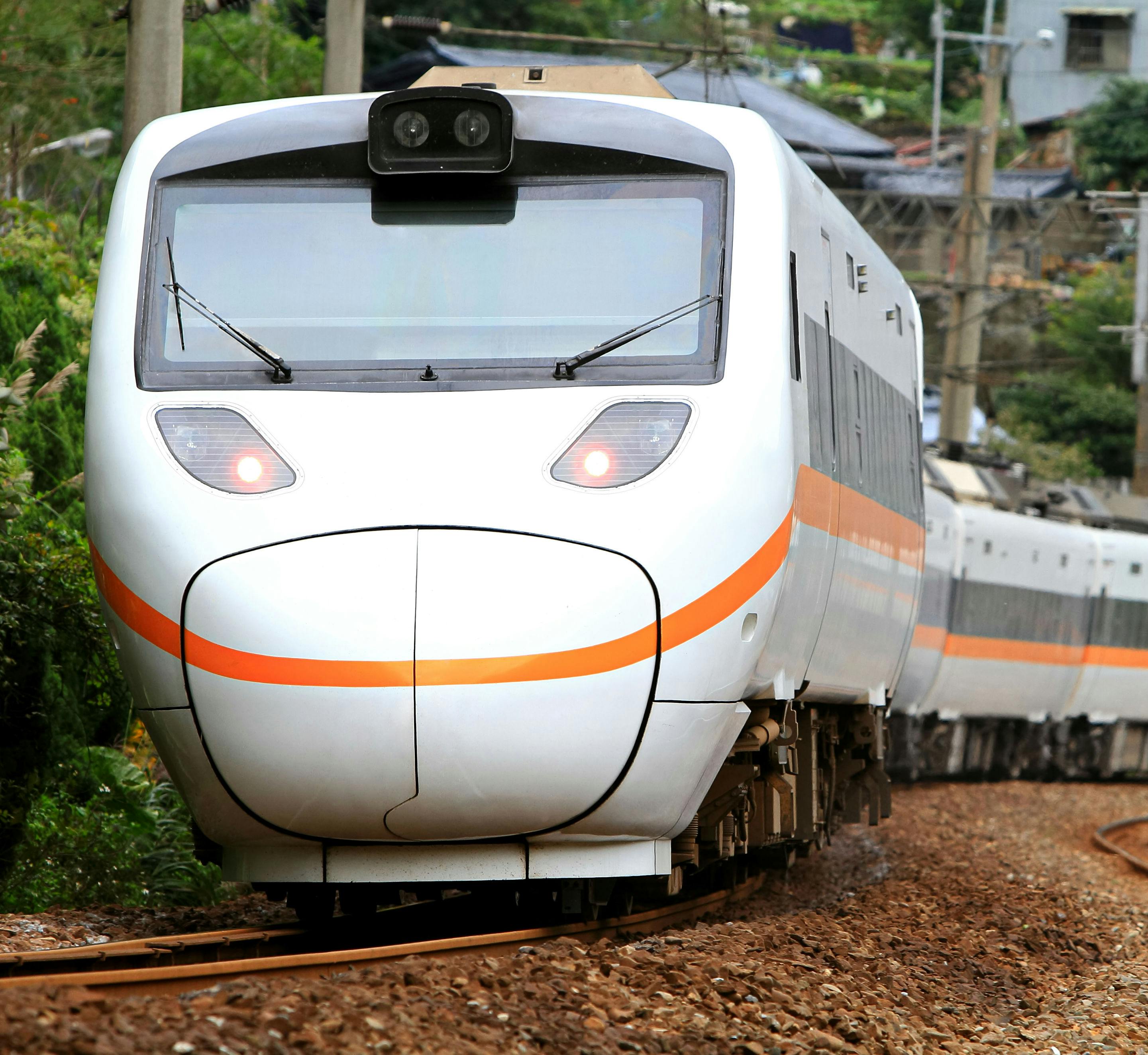 Ένα σύγχρονο λευκό τρένο με πορτοκαλί ρίγα ταξιδεύει σε μια διαδρομή μέσα από μια δασώδη περιοχή.