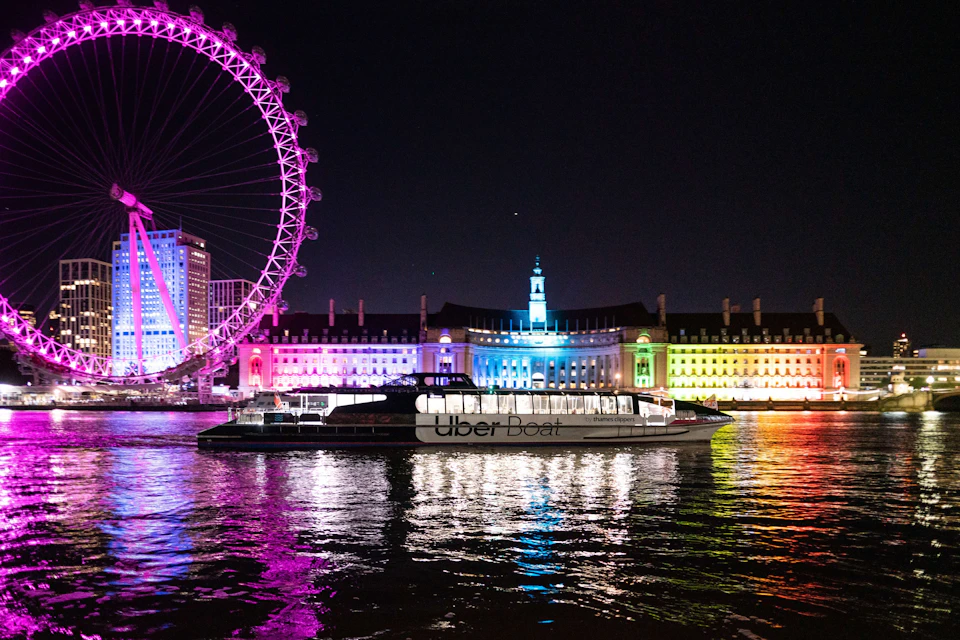 A nighttime view of the London Eye and a brightly lit building, with an Uber Boat on the Thames River in the foreground.