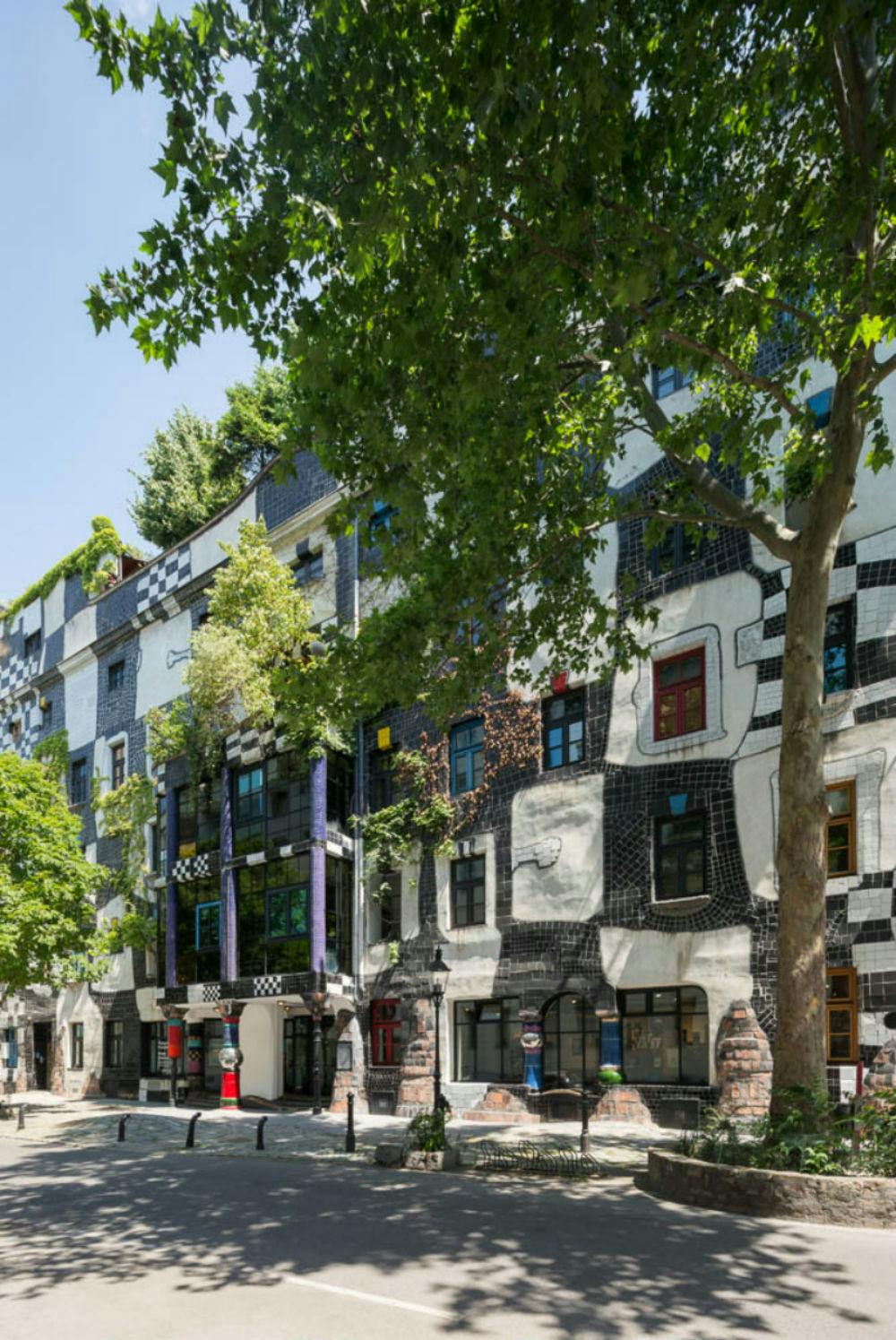 Красочный, эклектичный экстерьер здания с нерегулярными узорами, в окружении пышных зеленых деревьев, под чистым голубым небом.