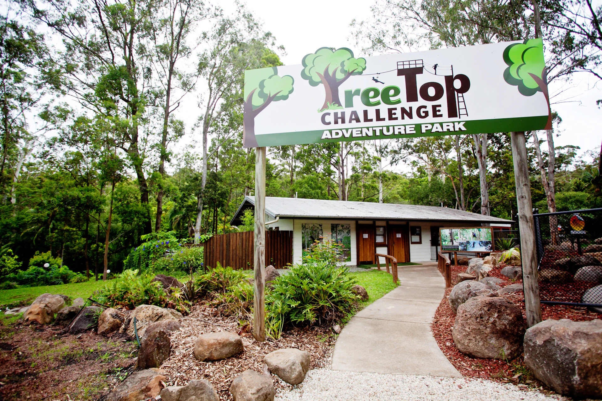 Entrance to TreeTop Challenge Adventure Park with a sign above a pathway leading to a small building surrounded by trees and rocks.
