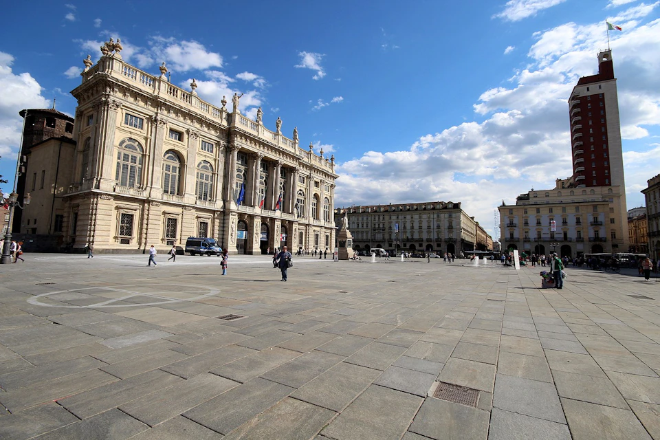 Large plaza with people walking, surrounded by ornate historic buildings under a partly cloudy blue sky.