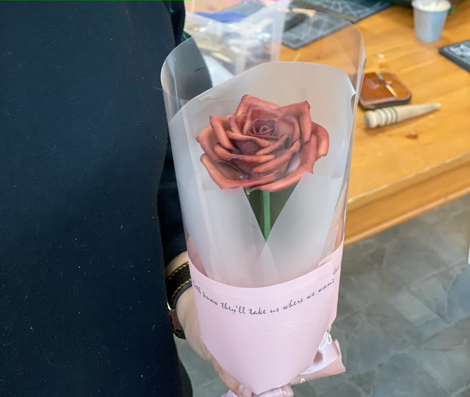A hand holding a single artificial rose wrapped in transparent and pink paper with text, placed in an indoor setting.