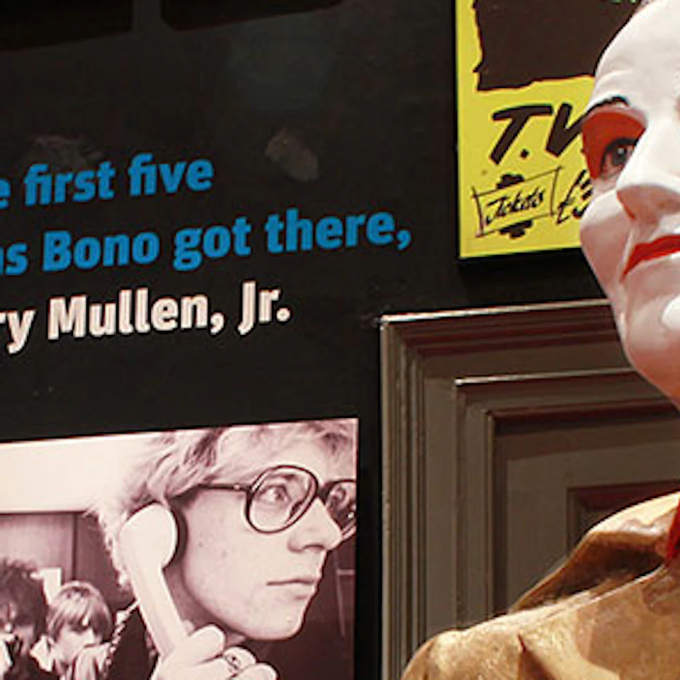 A museum display featuring memorabilia from Eagles, U2, and a statue of a man in a gold suit with red accents and red horns.