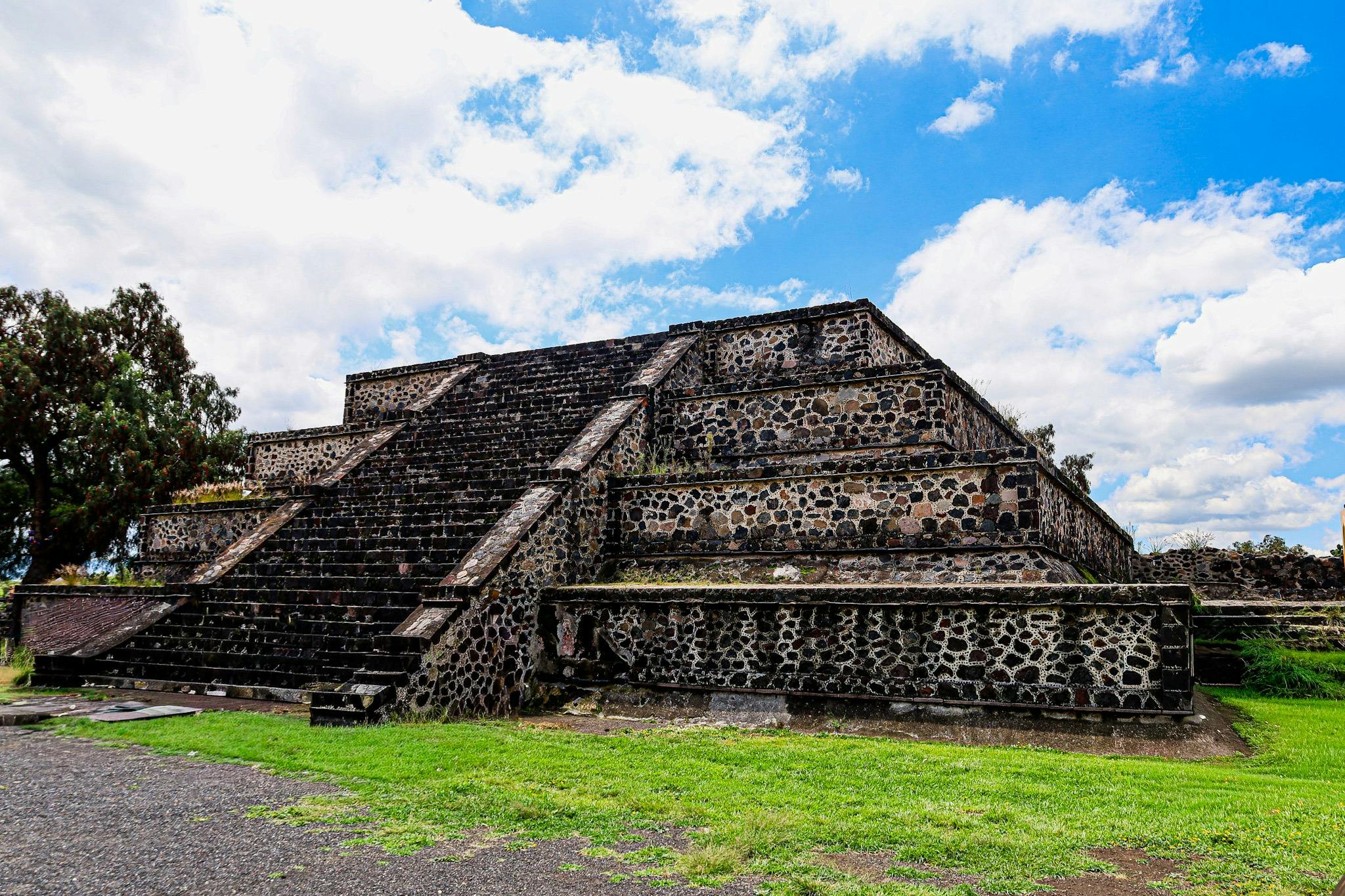 A stepped stone pyramid under a partly cloudy blue sky, surrounded by green grass and a paved area.