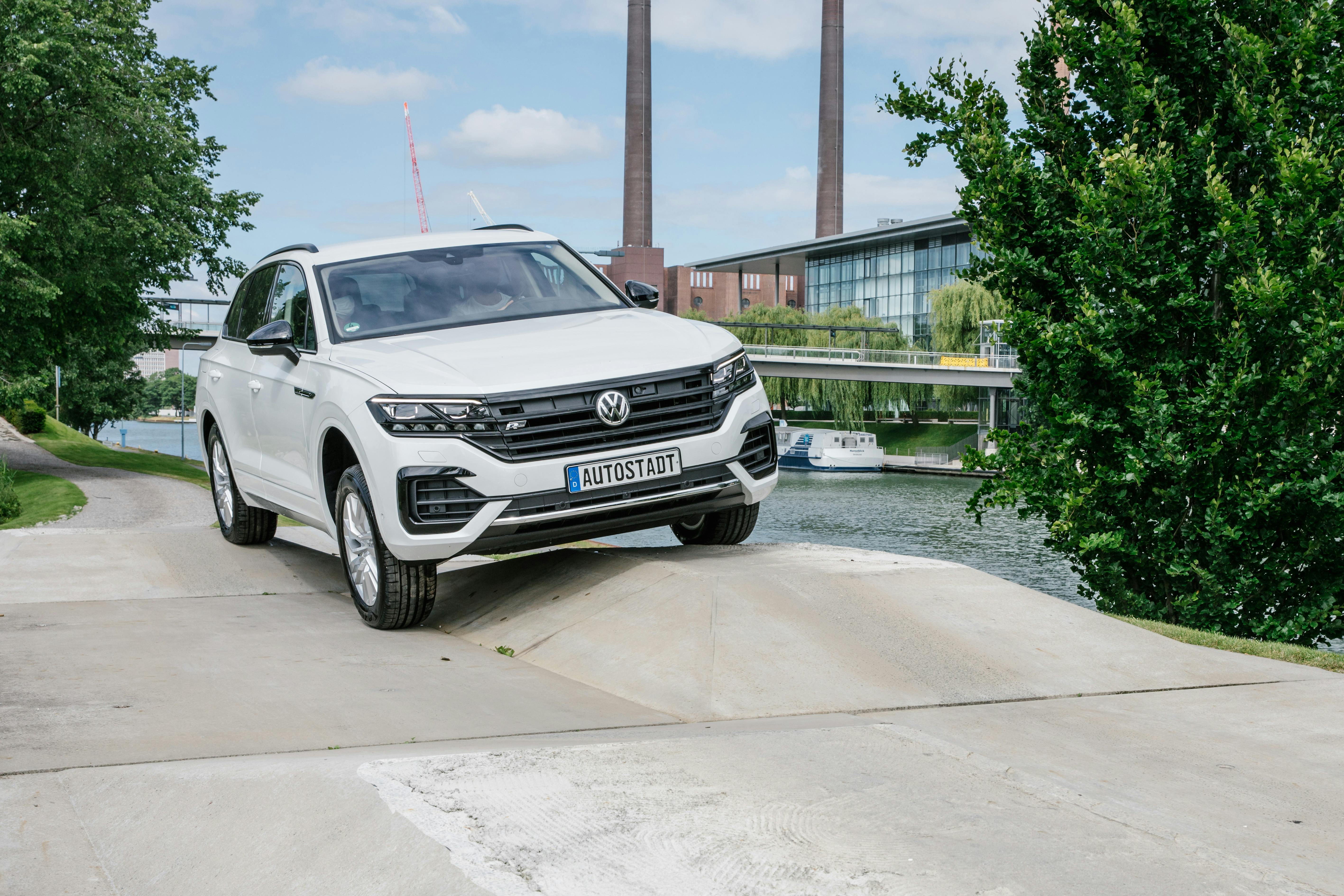 A white Volkswagen SUV on an inclined surface by a river, with industrial buildings, a bridge, and greenery in the background.