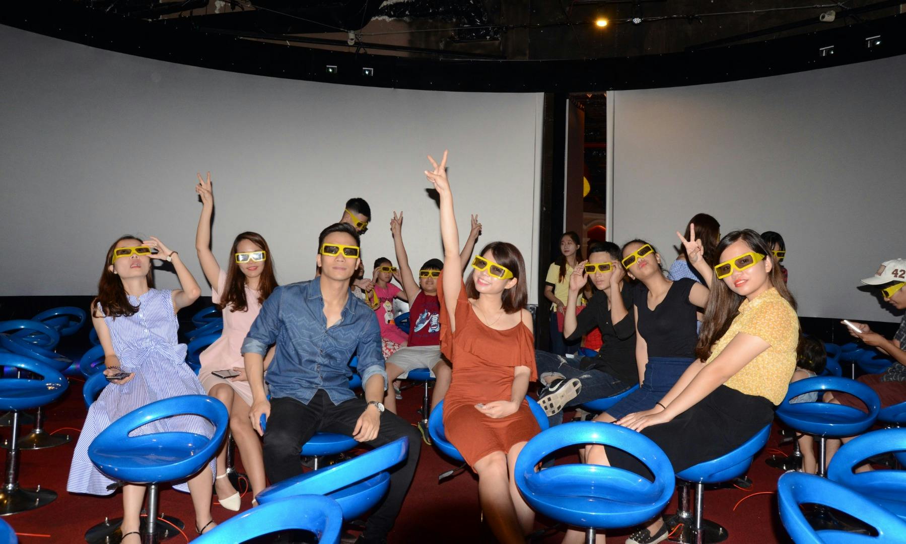 A group of people wearing 3D glasses sitting on blue stools in a theater-like setting, some raising their hands enthusiastically.