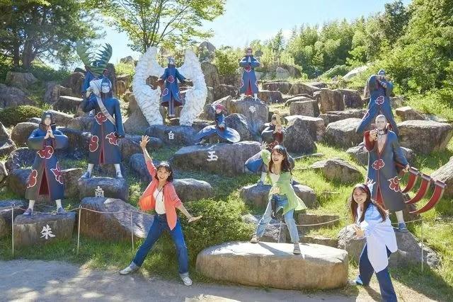 Three people posing enthusiastically in a park with statues of characters from an anime series among rocks and greenery.