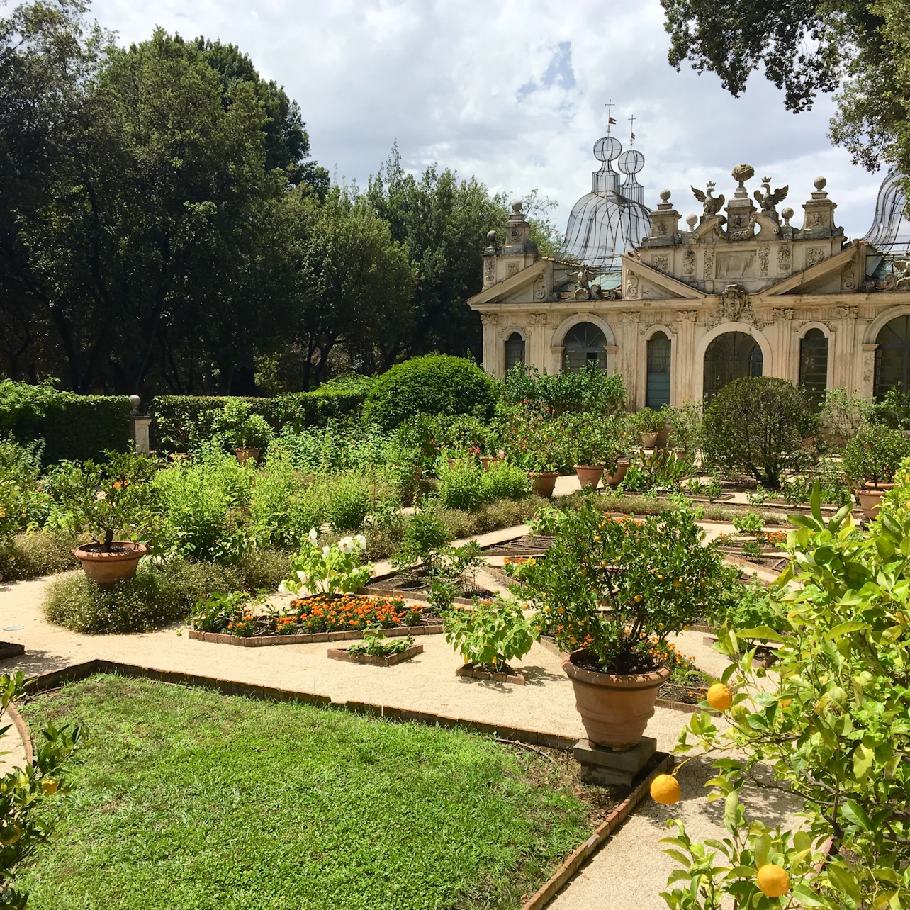 Lush garden with stone paths, various plants in pots, shrubs, and a historical building with ornate decorations in the background.