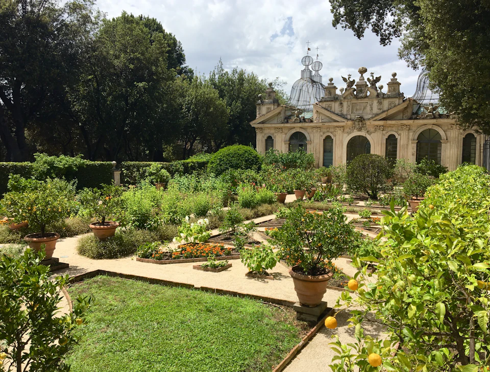 Lush garden with stone paths, various plants in pots, shrubs, and a historical building with ornate decorations in the background.