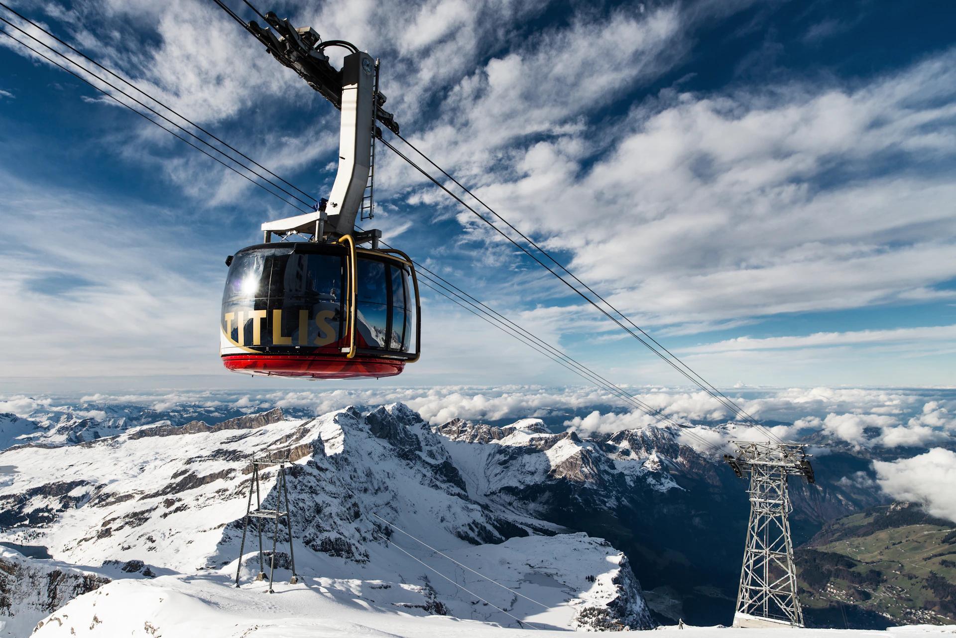 A cable car labeled "Titlis" suspended over snow-covered mountains, with a cloudy sky in the background.