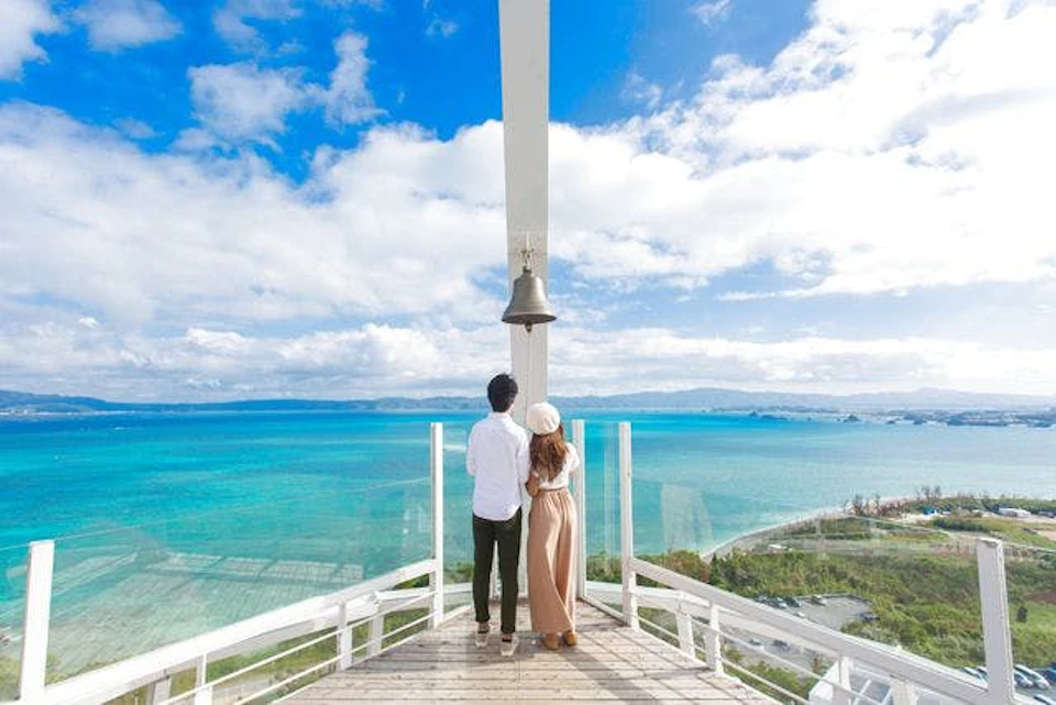 A couple standing on a wooden platform overlooking a turquoise ocean, with a large bell and bright blue sky in the background.